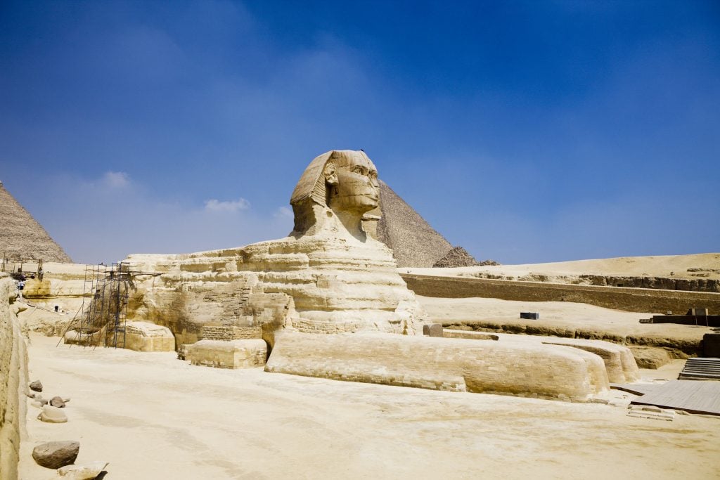 A photograph of the Great Sphinx of Giza depict against blue skies from the sculpture's right side