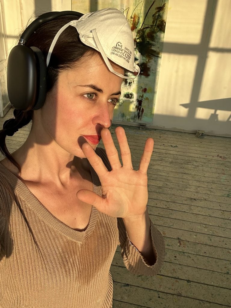 a woman in a selfie stands wearing earphones and a medical mask is on her head, she is holding up her palm to the camera