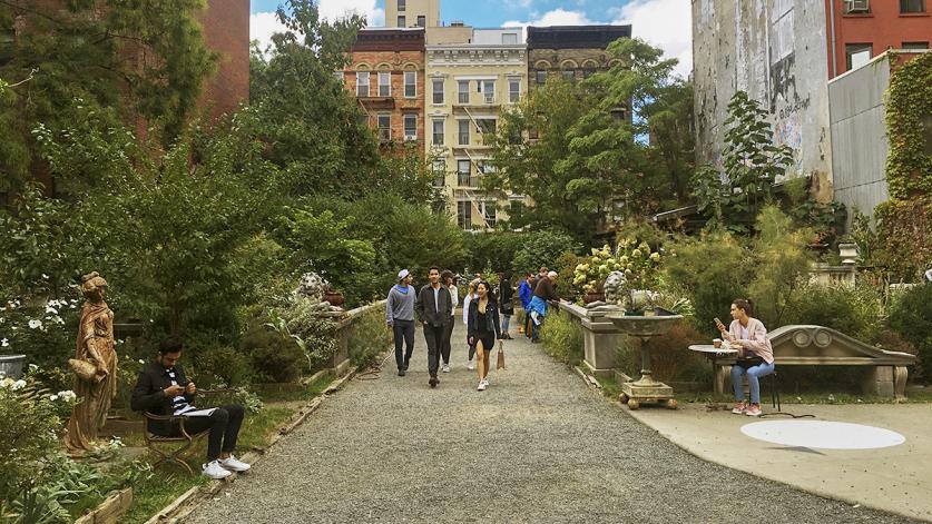 A group of people walk down a gravel path in the center of an urban garden, lined with trees, flowers, and a balustrade flanked by two lion statues.