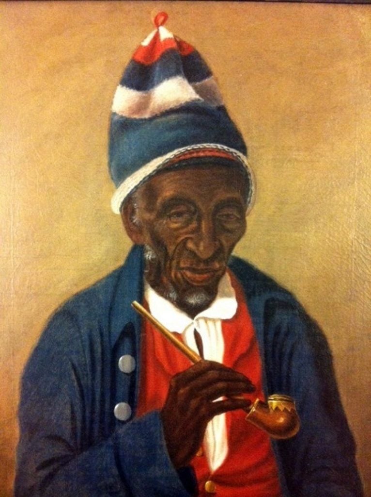 A traditional looking 19th-century oil painting of an elderly Black man, seen from the chest up in front of a warm hued background. He is wearing a kufi, a knit hat worn by African Muslim men, which is tall with large horizontal stripes in red, white, and blue. He also wears a blue collared jacket with silver buttons open to show a white collared shirt under a red sweater with gold buttons. He holds a long wooden pipe in his right hand.