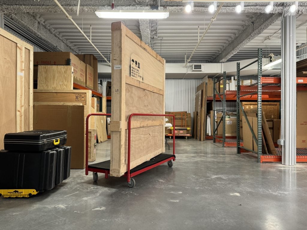 Inside a packing warehouse of Fine Art Shippers with a painting crate on a dolly and metal racks with cardboard boxes both full and unused.