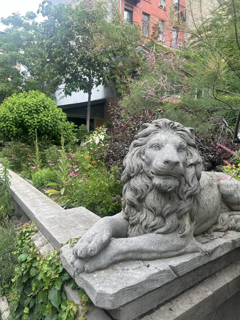 A stone lion statue perches at the end of a limestone balustrade in a verdant garden.