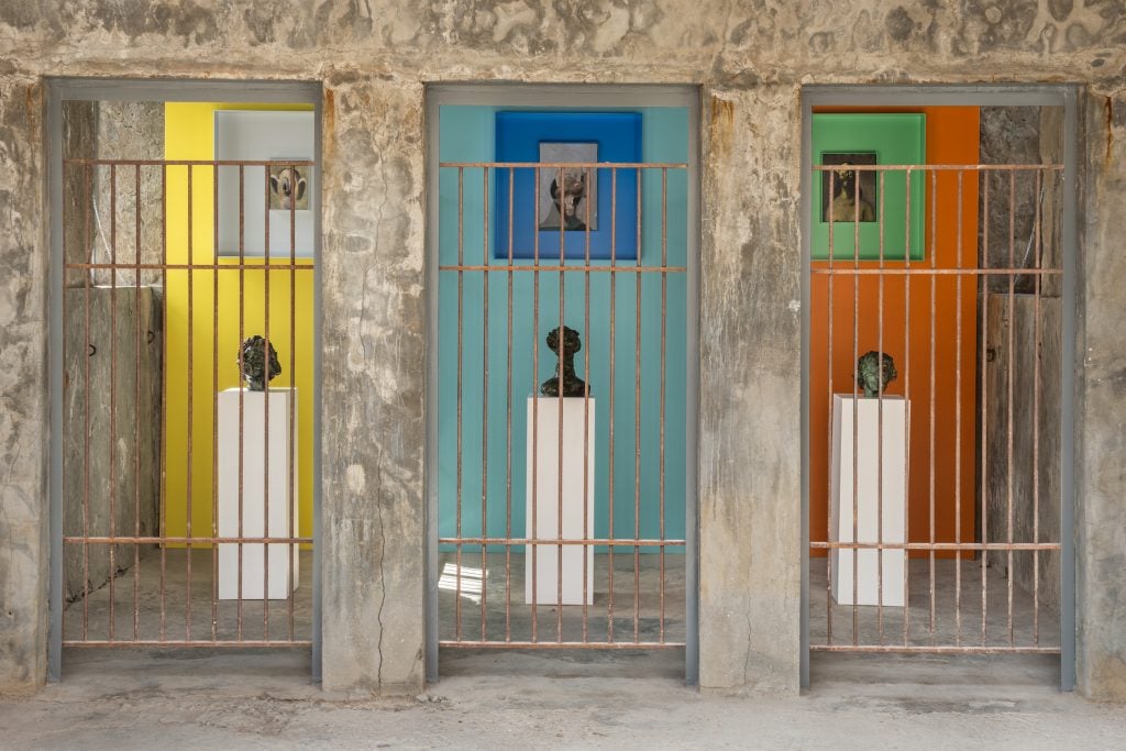 oil paintings and sculptures of heads on plinths are in three cages, with brightly colored painted walls in yellow, blue, and orange