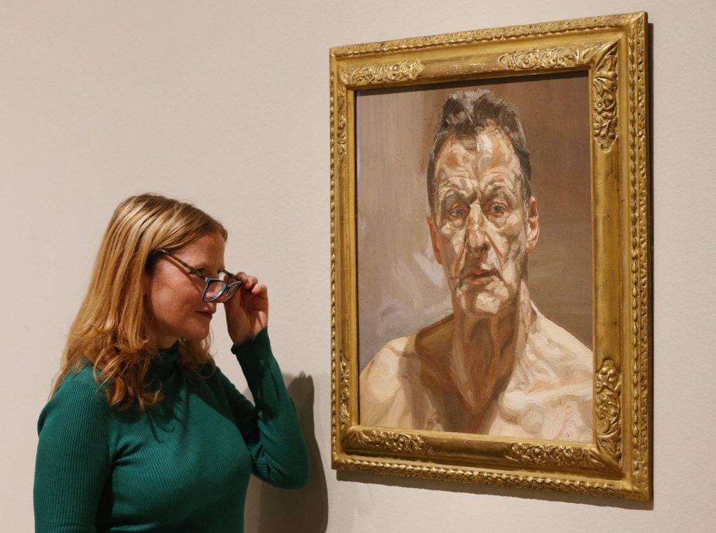 A blonde woman looks at Lucian Freud's portrait of a topless white men, painted in expressive strokes.