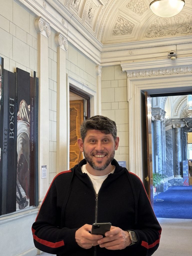 Portrait photo of Ilya Kushnirskiy of Fine Art Shippers standing in a grand New York apartment building foyer with the door open to the street behind him and some paintings installed on the walls that have molding. Wearing a black zip-up hoodie with one red stripe down each arm over a white shirt, holding his cellphone with both hands and smiling.