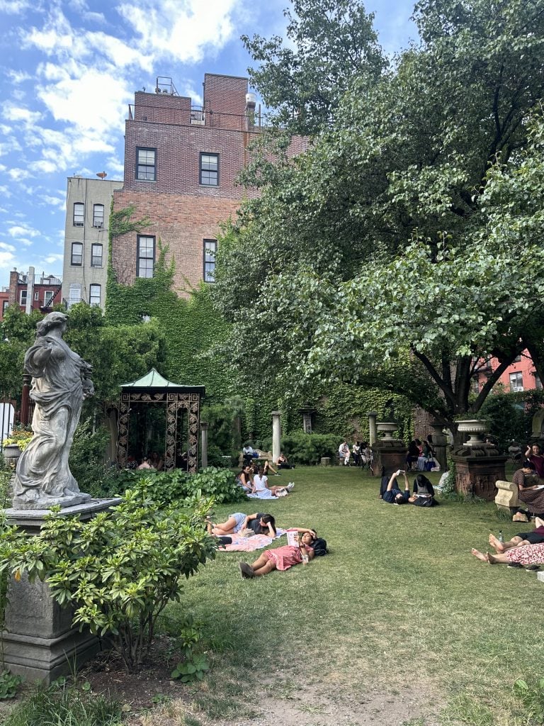 A large neoclassical statue of a woman in front of an ornate iron gazebo in an urban garden where visitor lie down on the grass lawn relaxing.