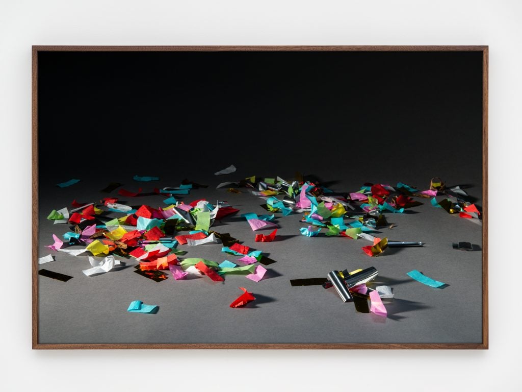 A photo by rose salane of a pile of multicolor paper confetti and paper strings on a grey ground and diffuse black background.