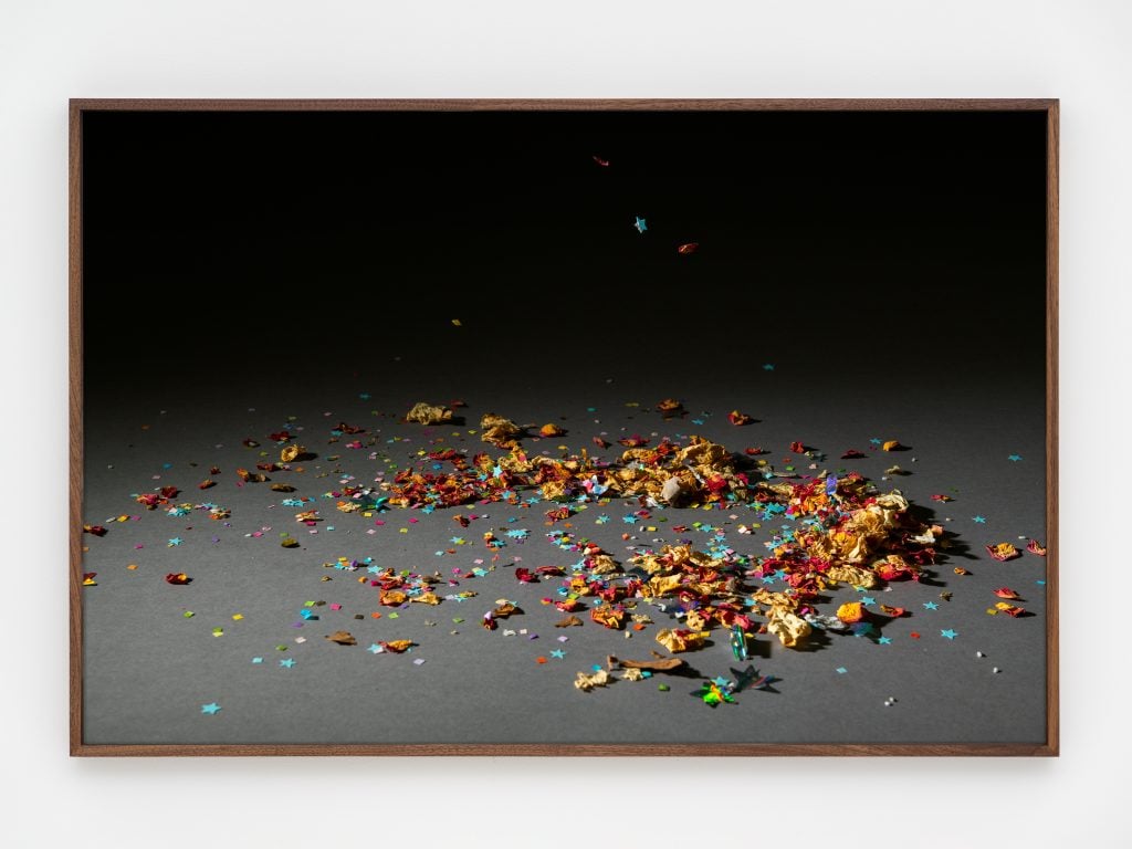 A photo by rose salane of a pile of multicolor paper confetti on a grey ground and diffuse black background.