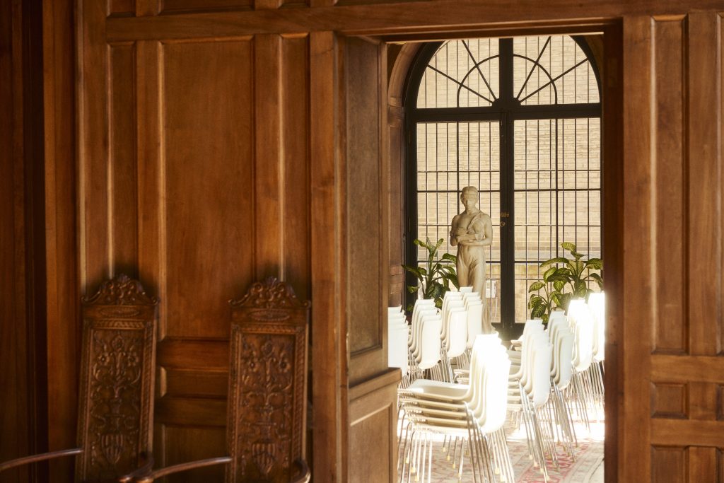 Entrance to an auditorium, white chairs stacked in front of a classical marble sculpture of a woman, two plants, and an arched glass door, seen through a wooden doorway. .