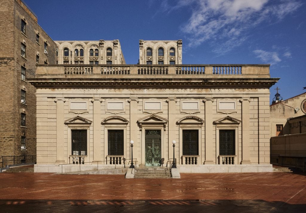 A Beaux Arts building with a brick courtyard in front of it, with a bright blue sky.